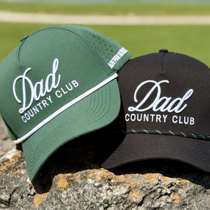 Two 'Dad Country Club' hats, one green, one black, on a rock with a blurred green field background